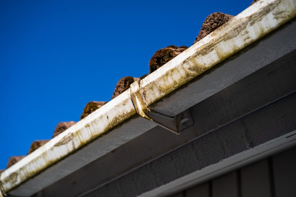 A person safely cleaning leaves and debris from a home’s rain gutter using gloves and a small scoop on a ladder.