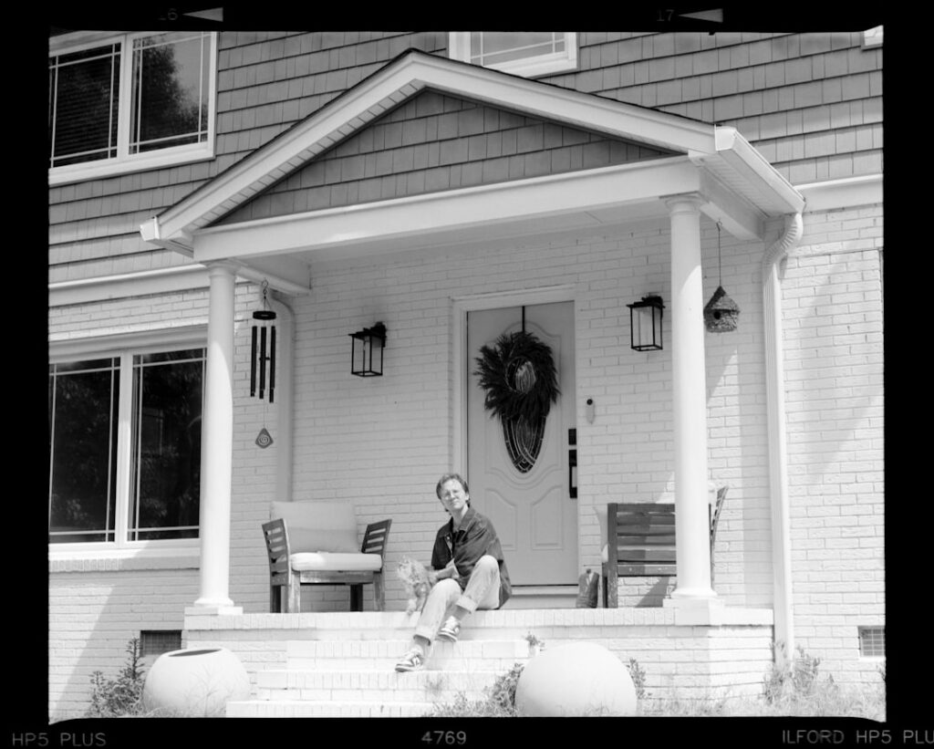 A craftsman-style front porch with railings, potted flowers, and seating on a sunny afternoon.
