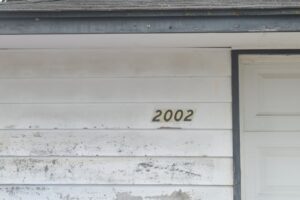 A homeowner in safety glasses using a power washer on vinyl siding with nearby garden plants protected by plastic.