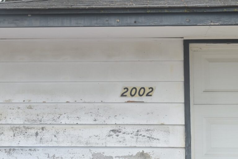 A homeowner in safety glasses using a power washer on vinyl siding with nearby garden plants protected by plastic.