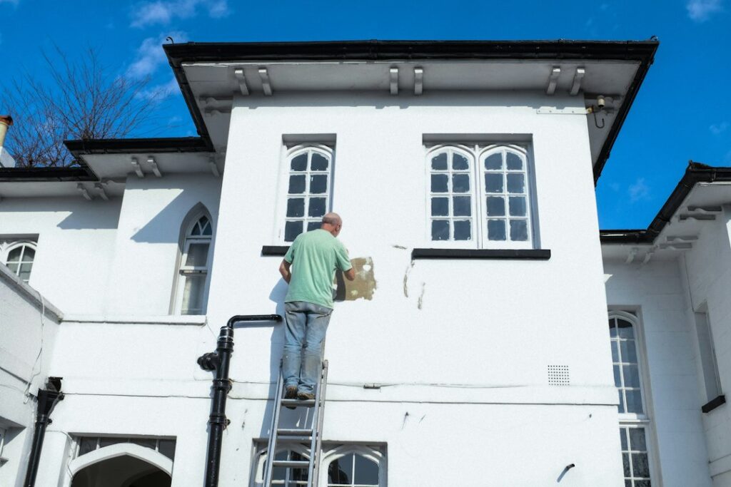 Homeowner painting house siding while another person steadies a ladder beside prepared drop cloths and tools.